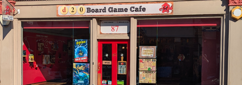 Front of "d20 Board Game Cafe" features a bright red door with posters, large windows displaying games, and warm, inviting lighting inside.