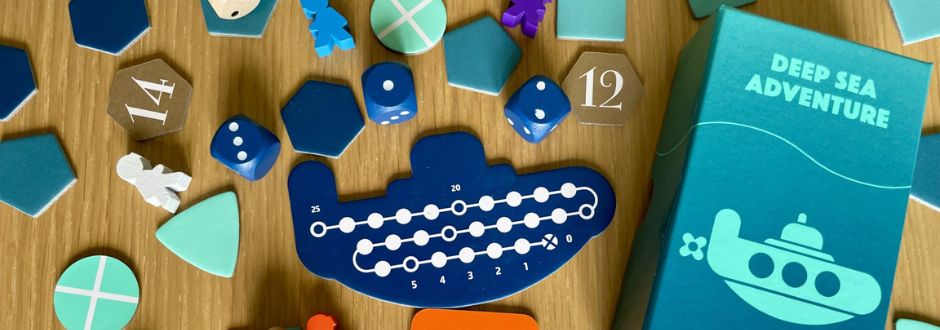 Board game components for 'Deep Sea Adventure' scattered on a wooden surface. Includes blue submarine, hexagonal pieces, dice, and a game box