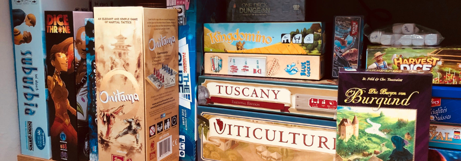 A shelf of stacked board games including Onitama, Kingdomino, Viticulture, and The Castles of Burgundy, with colourful boxes arranged side by side.
