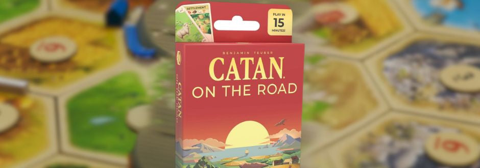  red box of 'Catan: On the Road' is displayed prominently, set against a blurred board game background. The box features a scenic illustration of a sunset over fields and mountains, with a playtime note of 15 minutes. 