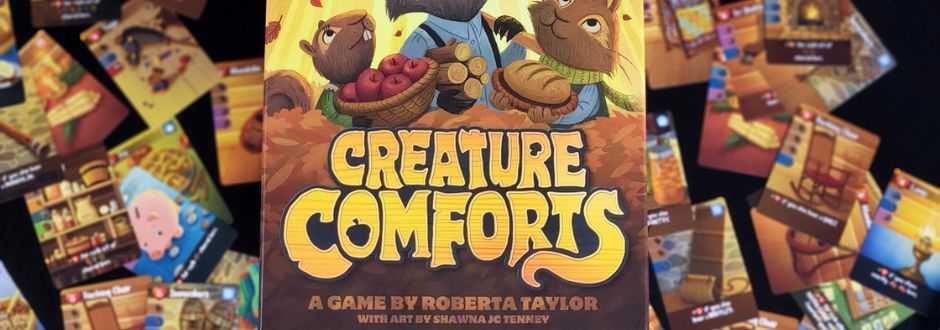 The image features the board game "Creature Comforts" by Roberta Taylor, with art by Shawna J.C. Tenney. The box shows cartoon animals holding autumn harvest items