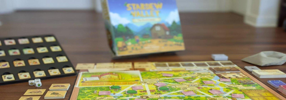A Stardew Valley board game setup on a table, with the village board, tiles, and components in the foreground and the game box blurred in the background.