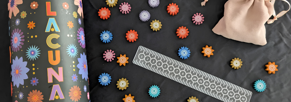 A colorful board game titled "Lacuna" is displayed, featuring vibrant floral pieces arranged on a black surface, a ruler, and a beige drawstring pouch.