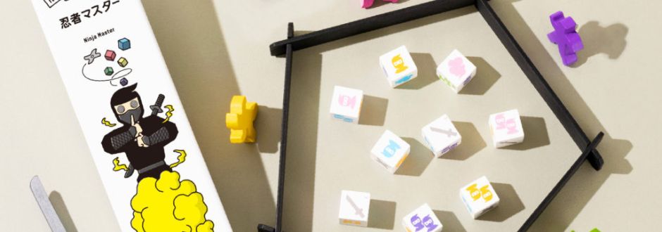 Board game setup with a white box featuring a cartoon ninja releasing colored falling blocks. Several multicolored cubes and small figures are scattered on a beige surface, creating a playful and strategic vibe.