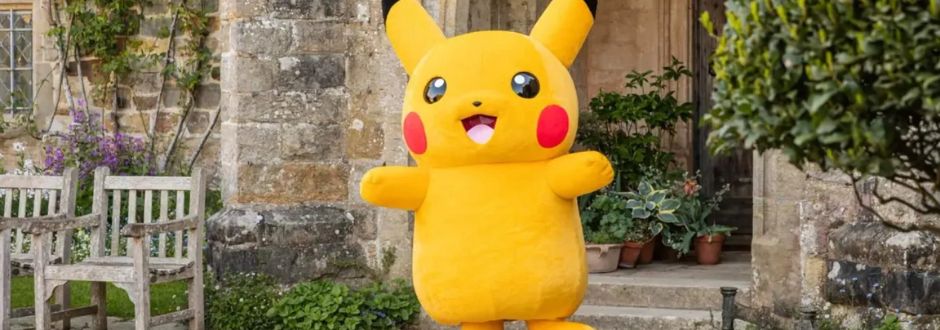 A large Pikachu mascot stands cheerfully in front of a rustic stone building with potted plants and wooden chairs, conveying a playful, welcoming vibe.