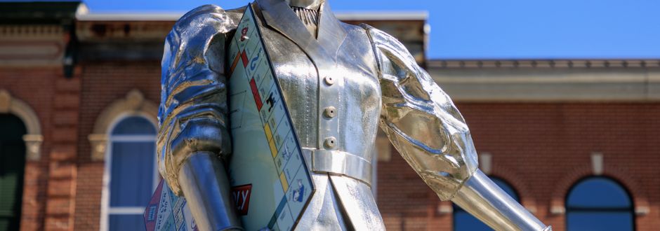 Close-up of a metallic statue shaped like a woman with puffy sleeves, wearing a sash made of Monopoly money. Set against a blue sky and brick building.