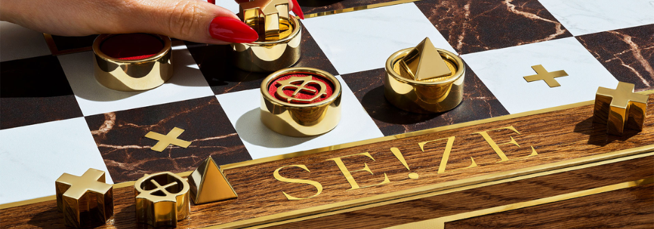 Close-up of a hand with red nails placing a gold game piece on a marble-and-wood board with geometric shapes and the word “SEIZE”.