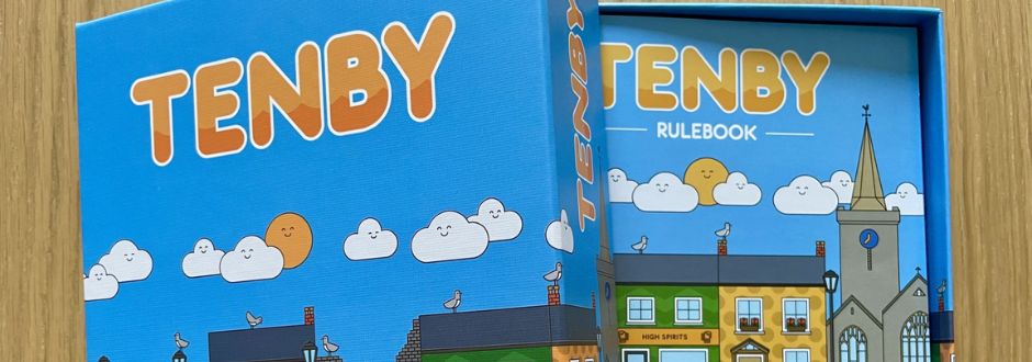A colorful board game box titled 'Tenby' with cartoon clouds and seagulls. The box lid is open, revealing a rulebook with a town illustration.