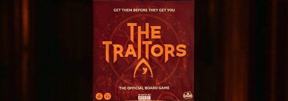 Box of "The Traitors" board game with bold orange text on a dark background, featuring a silhouette of a hooded figure. Tagline: "Get them before they get you."