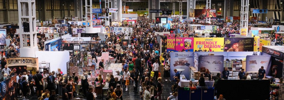 Crowded convention hall filled with booths, banners, and large groups of people browsing exhibits and games.