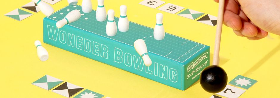 A mini bowling game on a yellow surface, featuring a turquoise box labeled "Wonder Bowling" with small white pins and a black ball on a stick. Surrounding cards add a playful touch.