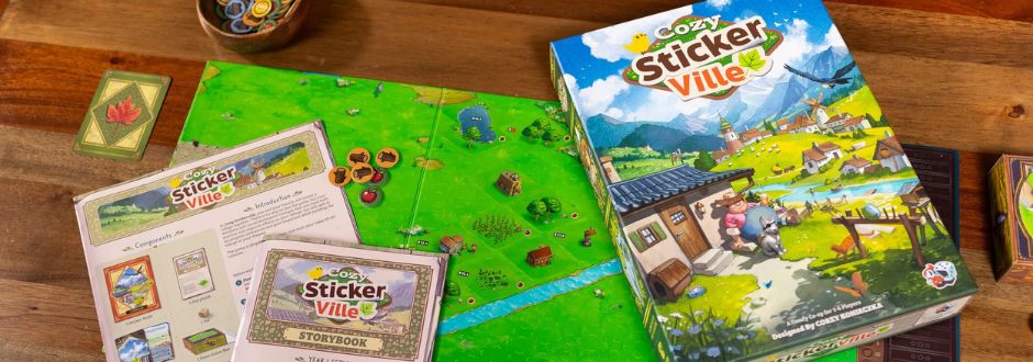 Box of "Cozy Stickerville" board game on a table, with colorful countryside artwork. Included are game components and instructions, evoking a playful, inviting atmosphere.