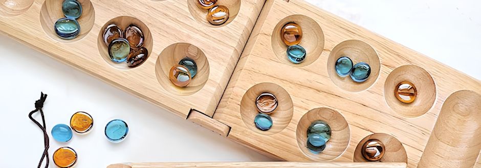 Wooden mancala game board with blue and amber glass stones in various wells. A black drawstring pouch with additional stones is nearby on a white surface.