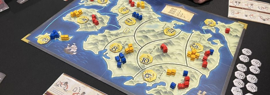A strategy board game in play features a map with numbered hexagonal regions and wooden pieces in red, blue, and yellow. Score tokens are nearby