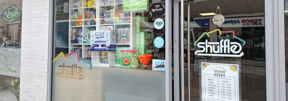 Shopfront of "Shuffle," featuring a large glass window displaying colorful board games. A sign with opening hours is visible. Tone is welcoming and playful.